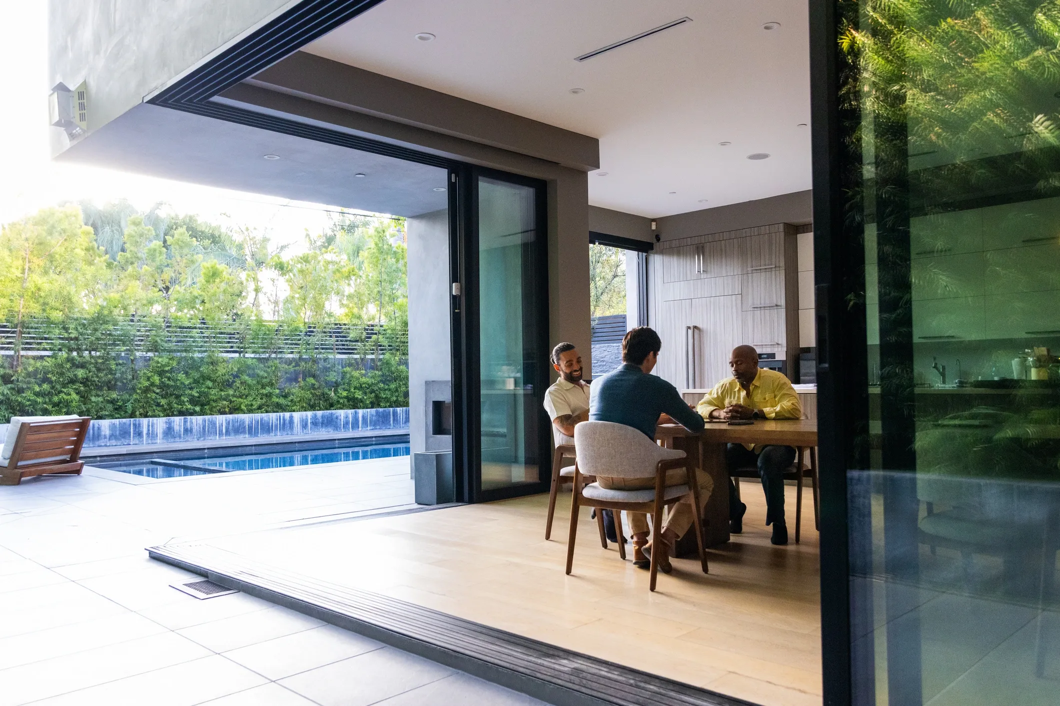 Three men sitting around a table in a room with a glass wall, a swimming pool, a bench