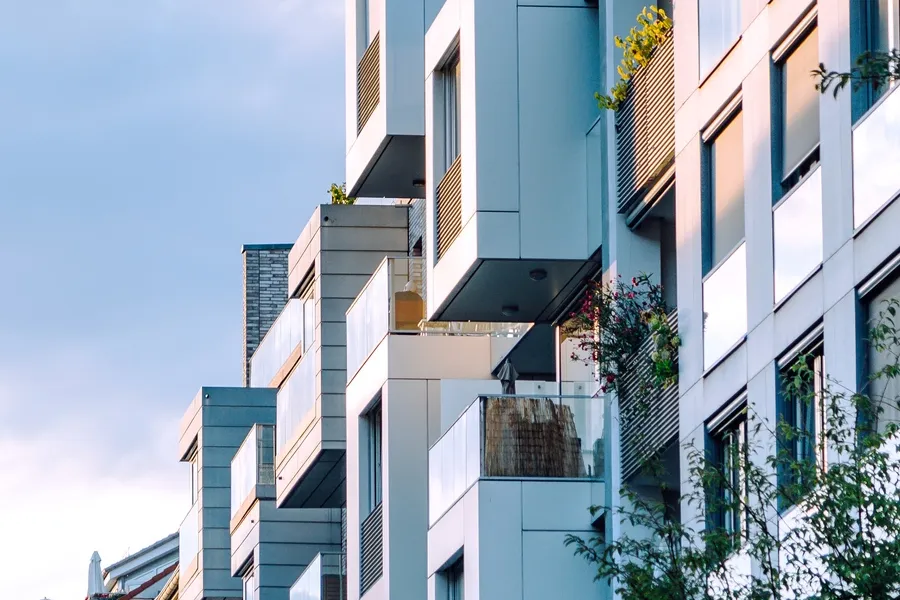 Tall building with many windows and balconies, surrounded by trees and plants, with a clear blue sky in the background