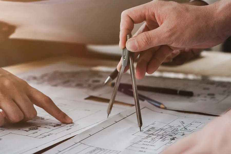 Two people working on a project with blueprints, one of them holding a compass and the other holding a pen