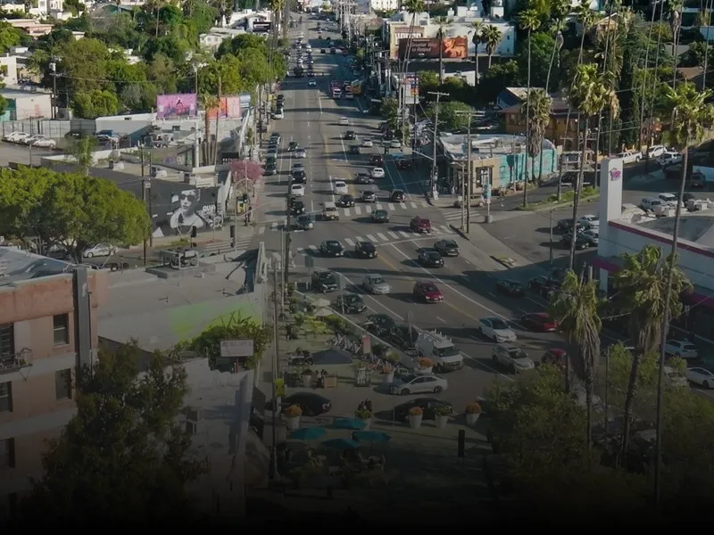 City street from the top of a building, with vehicles on the road, trees, buildings, poles, boards, hoardings
